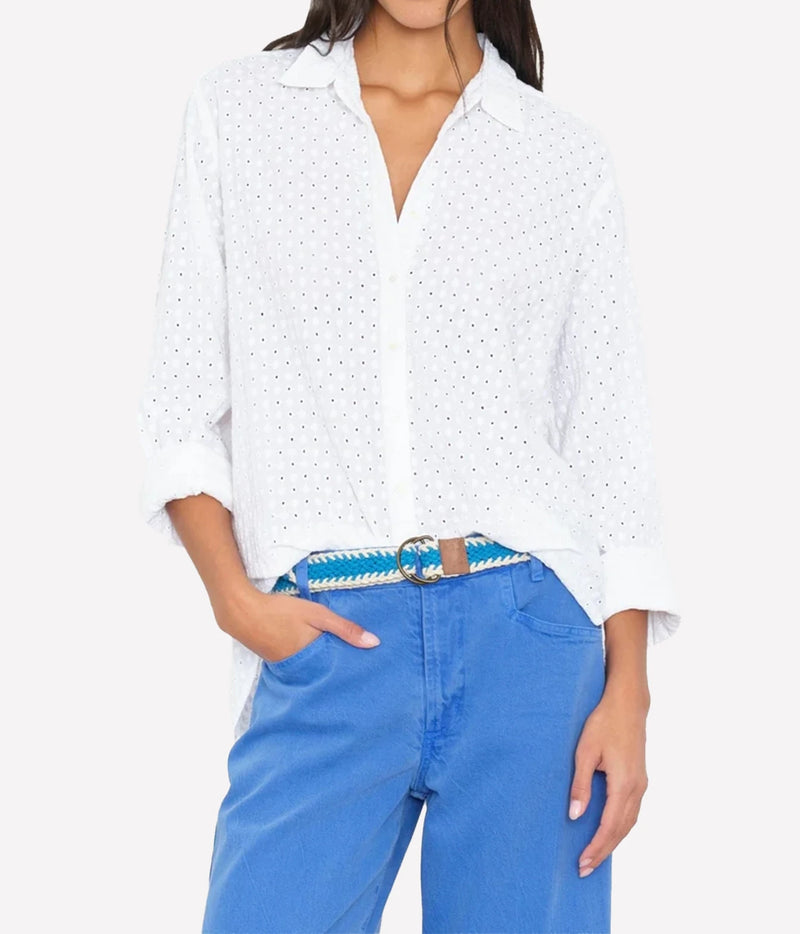 A woman wearing a white long-sleeve button-up shirt with a small eyelet pattern, tucked into blue jeans with a blue braided belt.