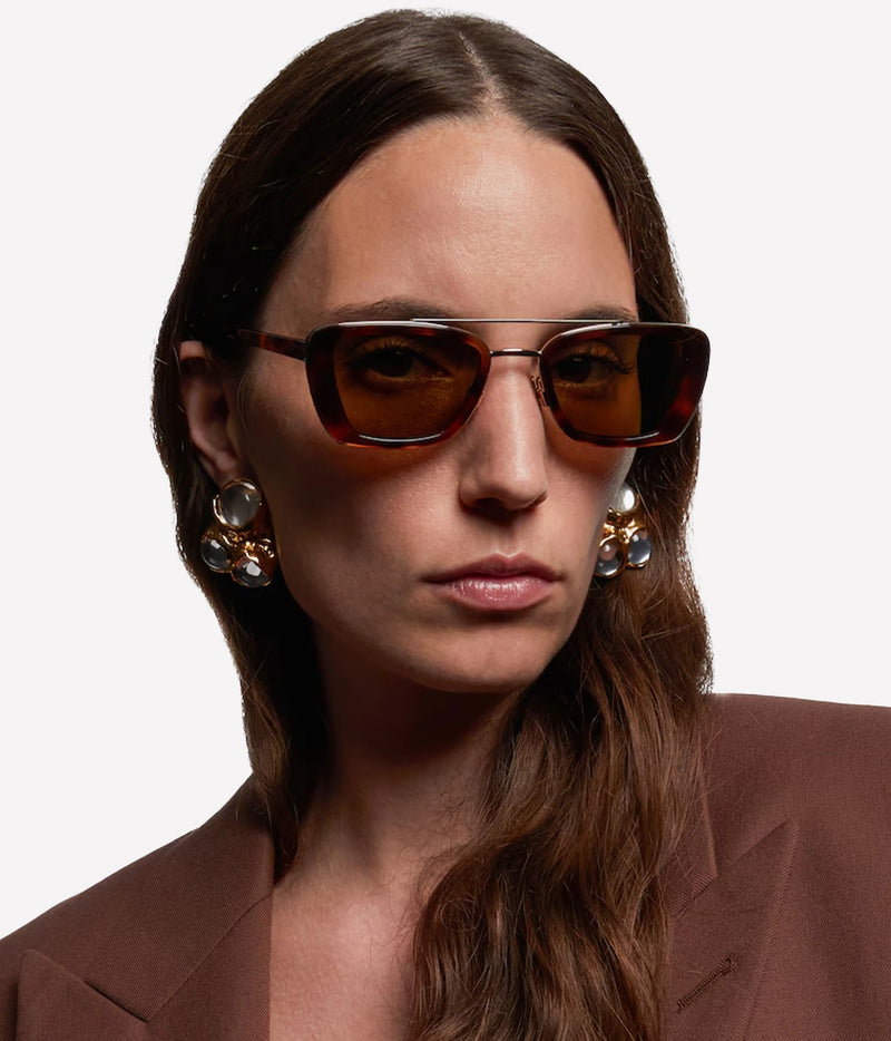 Close-up portrait of a woman wearing Saint Laurent Erin Sunglasses in Havana with a tortoiseshell frame, gold-tone metal detailing and brown tinted lenses, styled with statement earrings and a brown blazer.