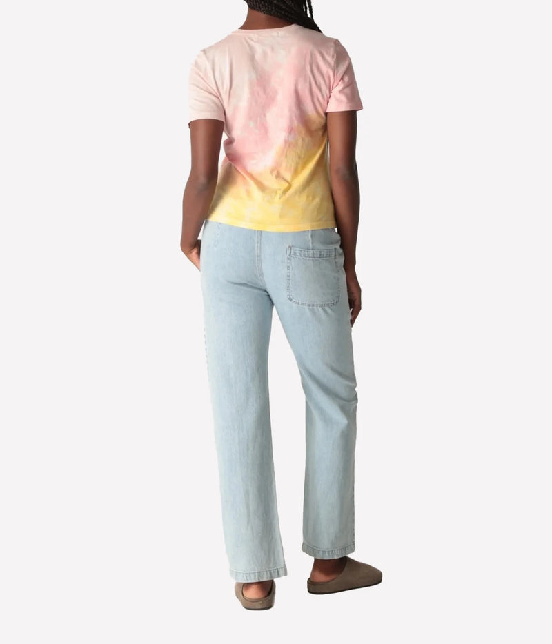 Back view of a model wearing the Electric Rose Everyday Haze T-Shirt in Sunrise Multi, showing the pink-to-yellow fade across the back, with light-wash straight-leg denim pants and taupe suede clogs