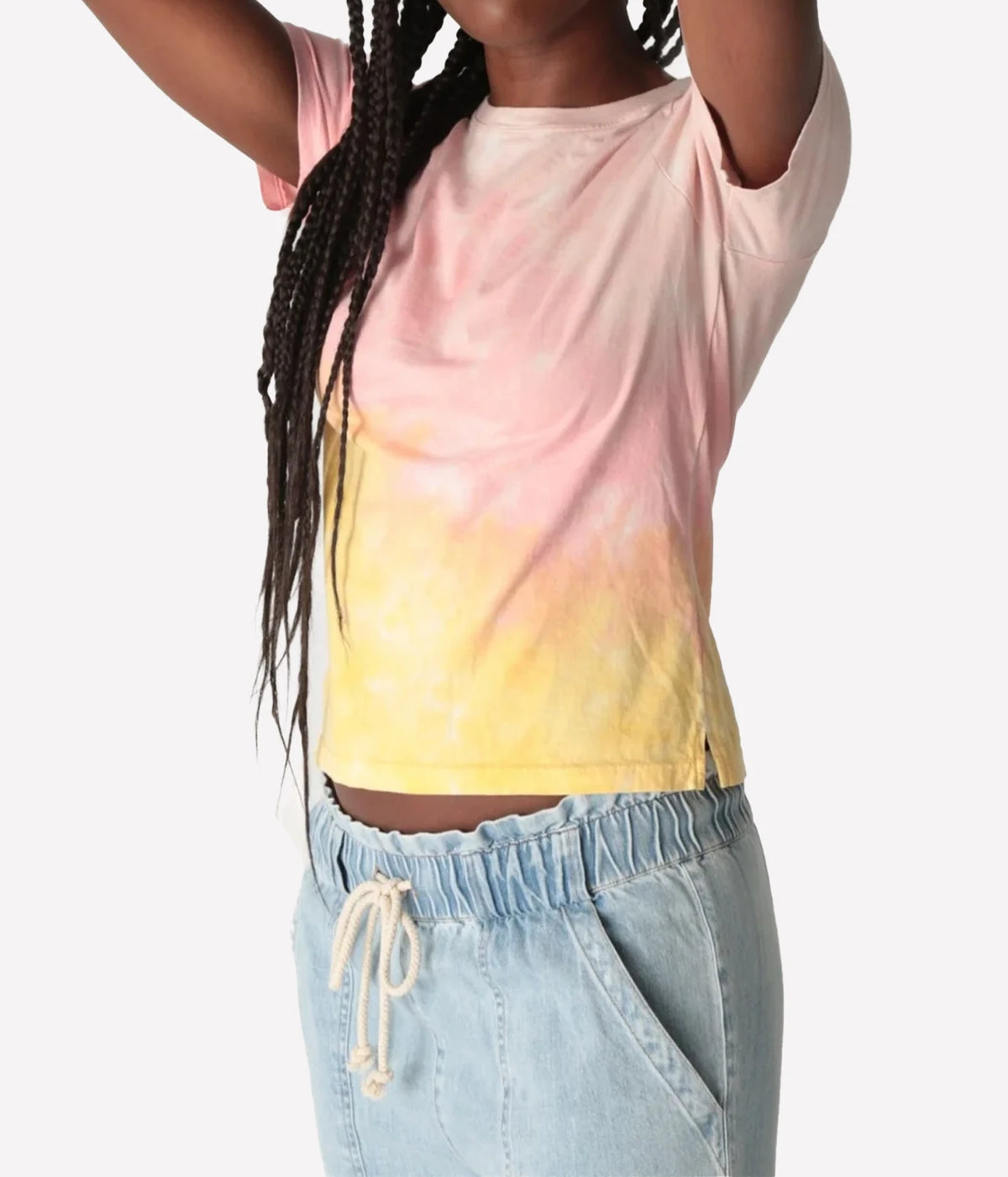 Close-up of a model with arms raised wearing the Electric Rose Everyday Haze T-Shirt in Sunrise Multi, showing the pink-to-yellow dip-dye, crew neck, short sleeves and split side hem, with light-wash denim drawstring pants