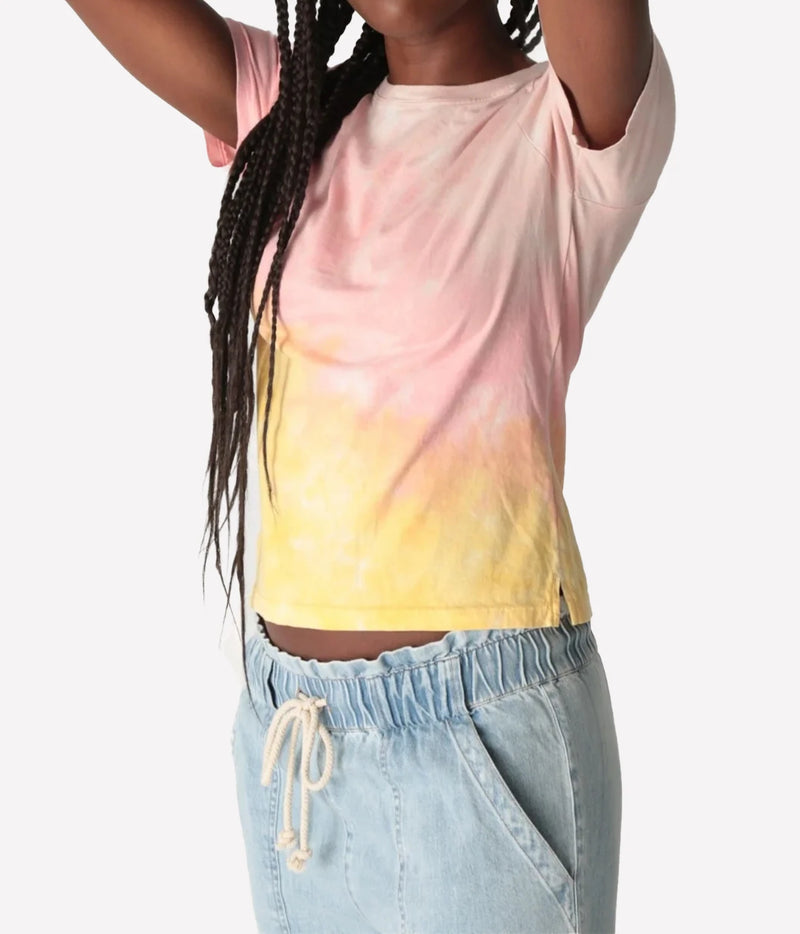 Close-up of a model with arms raised wearing the Electric Rose Everyday Haze T-Shirt in Sunrise Multi, showing the pink-to-yellow dip-dye, crew neck, short sleeves and split side hem, with light-wash denim drawstring pants