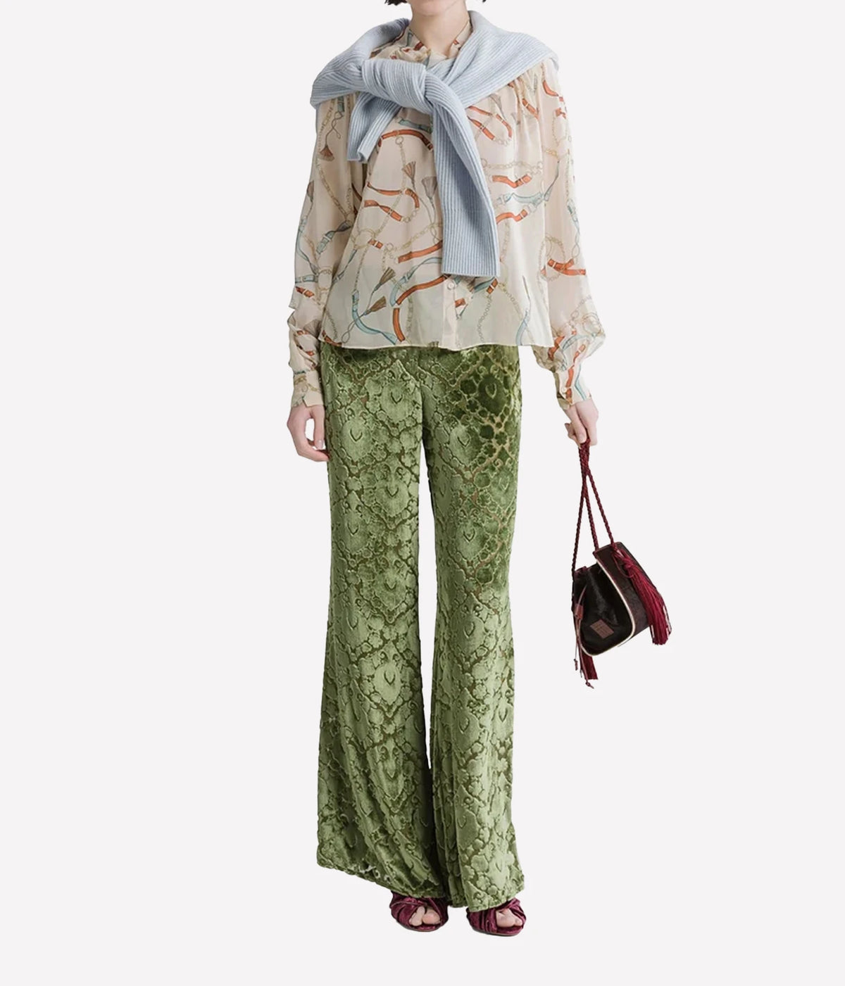 Woman wearing the Forte Forte crown print silk shirt in Ivory with green trousers, a blue knit around the shoulders and a dark handbag, full-length front view.