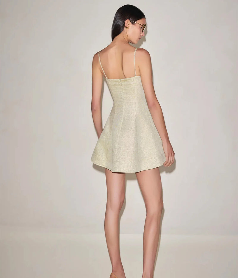 Full-length back view of a model wearing the Alexis Josefin Dress In Gold Mist with her face turned over her shoulder, showing the braided straps, fitted back and flared mini skirt