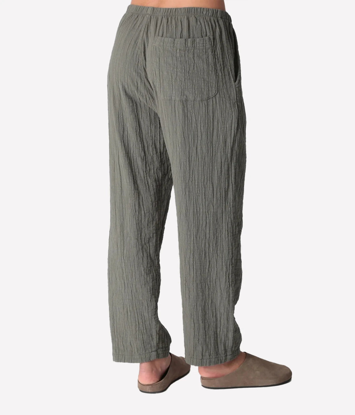 Back view of a model wearing the Electric Rose Kick Back Pant in Moss, showing the patch back pocket, elastic waist and relaxed fit, with taupe suede clogs