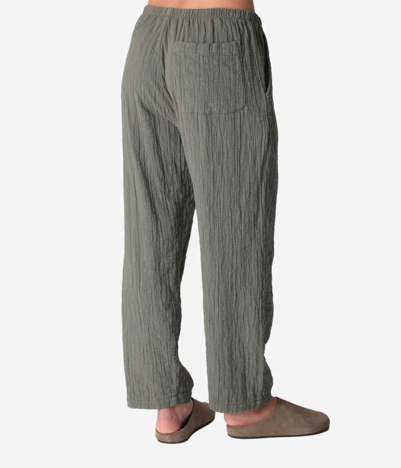 Back view of a model wearing the Electric Rose Kick Back Pant in Moss, showing the patch back pocket, elastic waist and relaxed fit, with taupe suede clogs