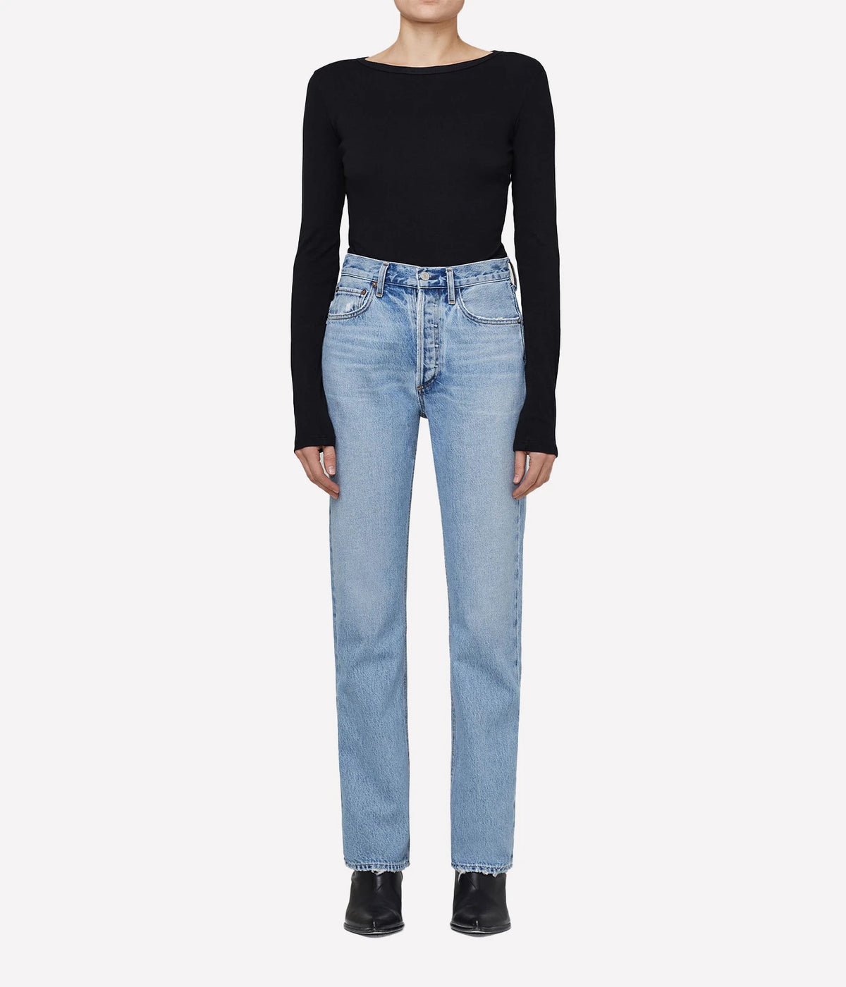 A model wearing a black long-sleeve boat-neck top and light-wash blue straight-leg jeans, paired with black pointed-toe heels, against a plain light gray background.