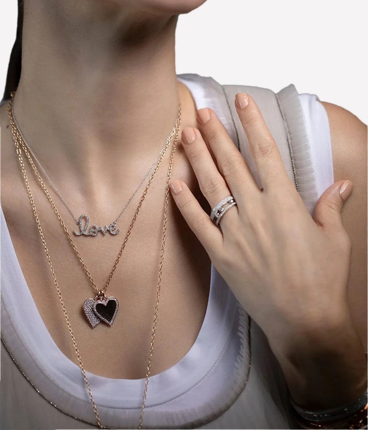 Model wearing the Sydney Evan Love Script Necklace In White Gold layered with a fine chain holding a dark heart pendant, with a white scoop-neck top and a diamond ring