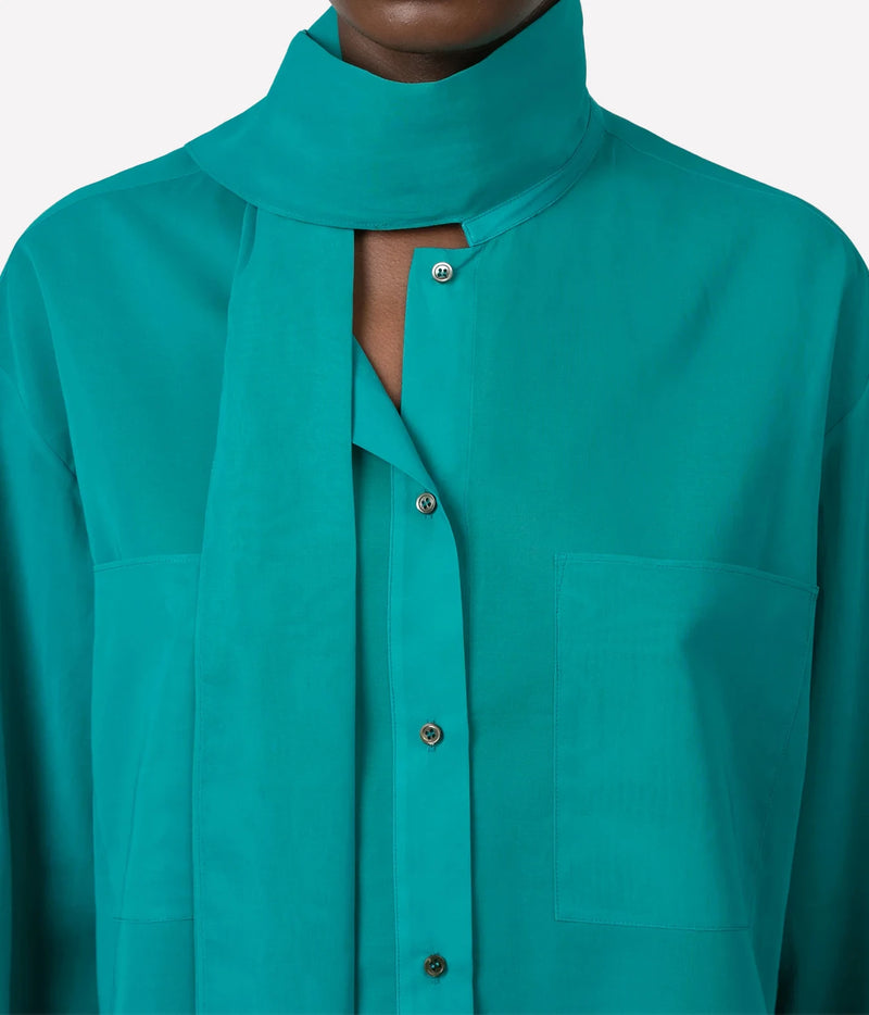A model wearing a vibrant teal long-sleeve blouse with a high ruffled collar and dark charcoal wide-leg trousers, shown from the neck down on a light gray background.