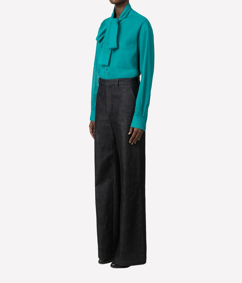 A model wearing a vibrant teal long-sleeve blouse with a high ruffled collar and dark charcoal wide-leg trousers, shown from the neck down on a light gray background.