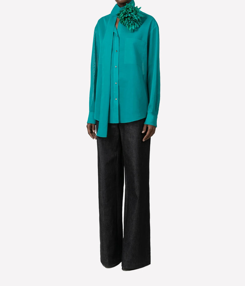 A model wearing a vibrant turquoise long-sleeve button-up shirt, black wide-leg trousers, and a large green textured flower collar accessory, set against a plain light gray background.