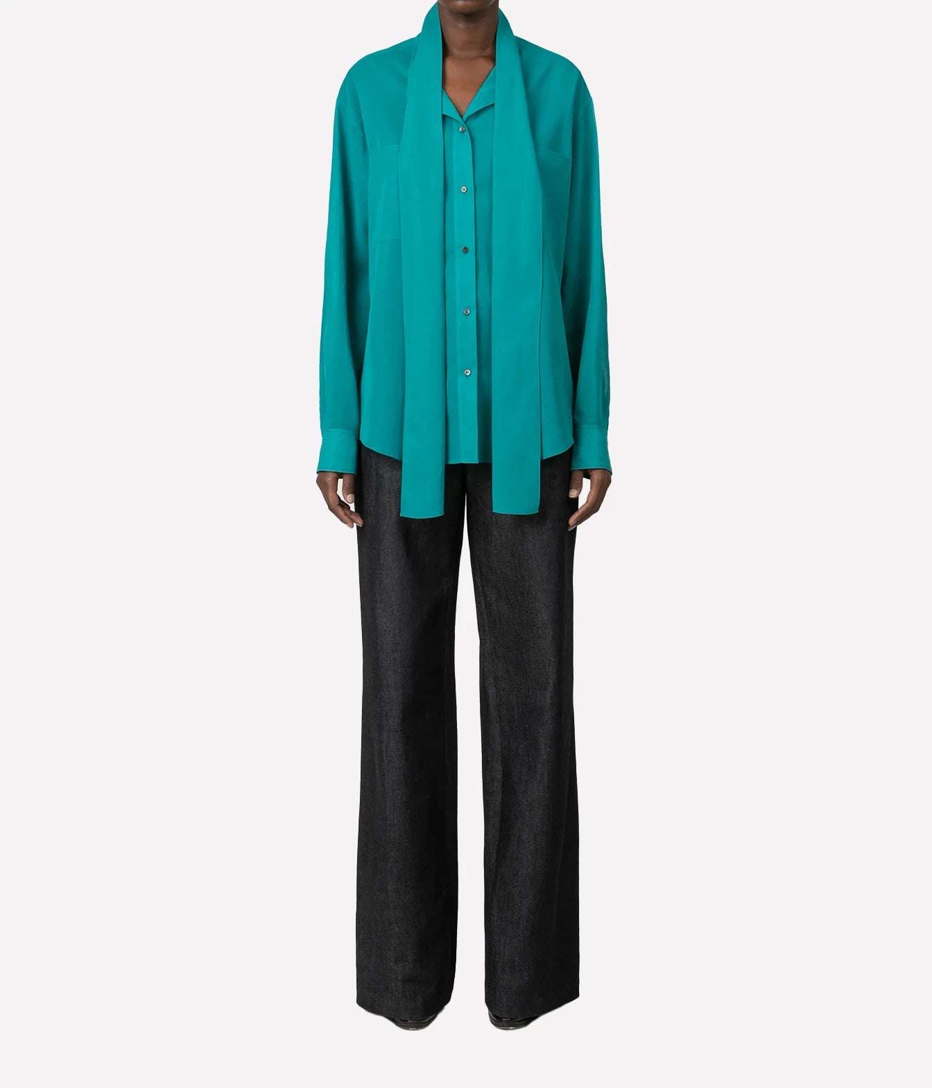 A model wearing a vibrant teal long-sleeve button-up shirt with a draped collar and dark charcoal wide-leg trousers, shot against a solid light gray background.