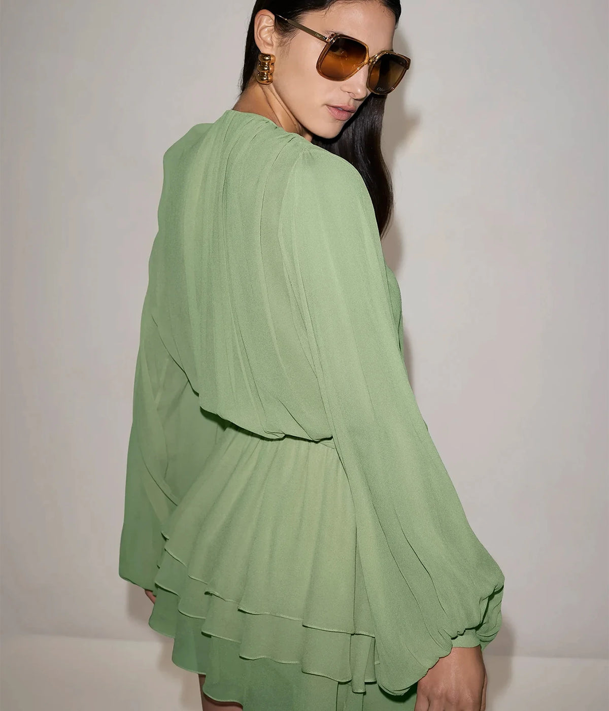Three-quarter view of a model wearing the Alexis Milosia Dress In Pistachio with amber sunglasses, showing the full volume of the long sleeves and the tiered mini skirt