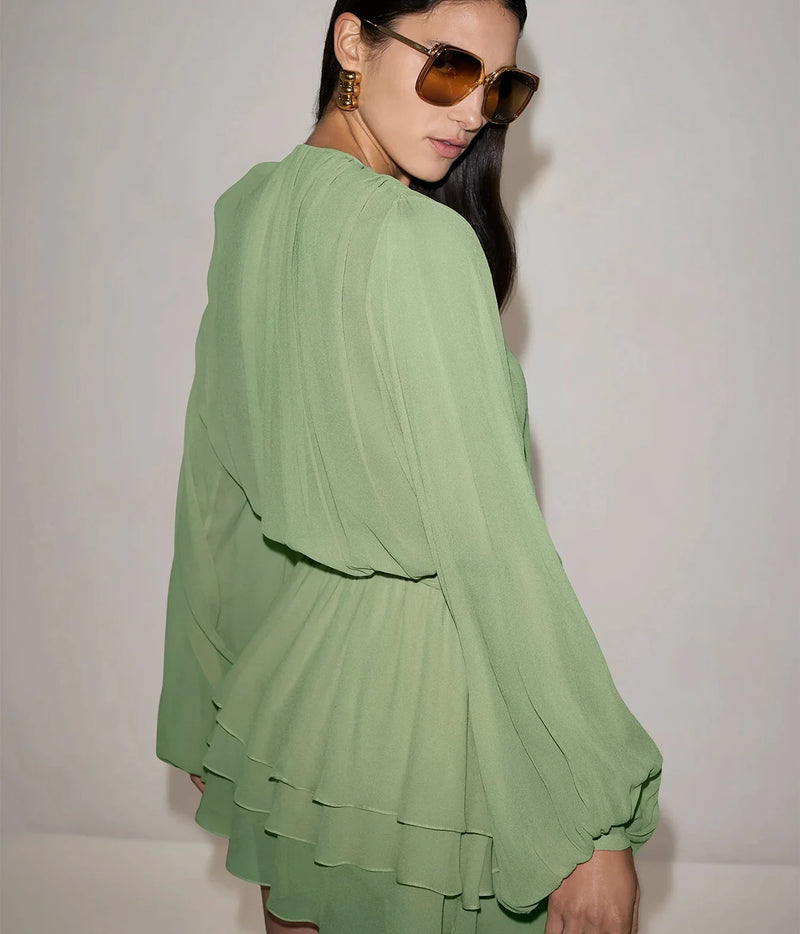 Three-quarter view of a model wearing the Alexis Milosia Dress In Pistachio with amber sunglasses, showing the full volume of the long sleeves and the tiered mini skirt