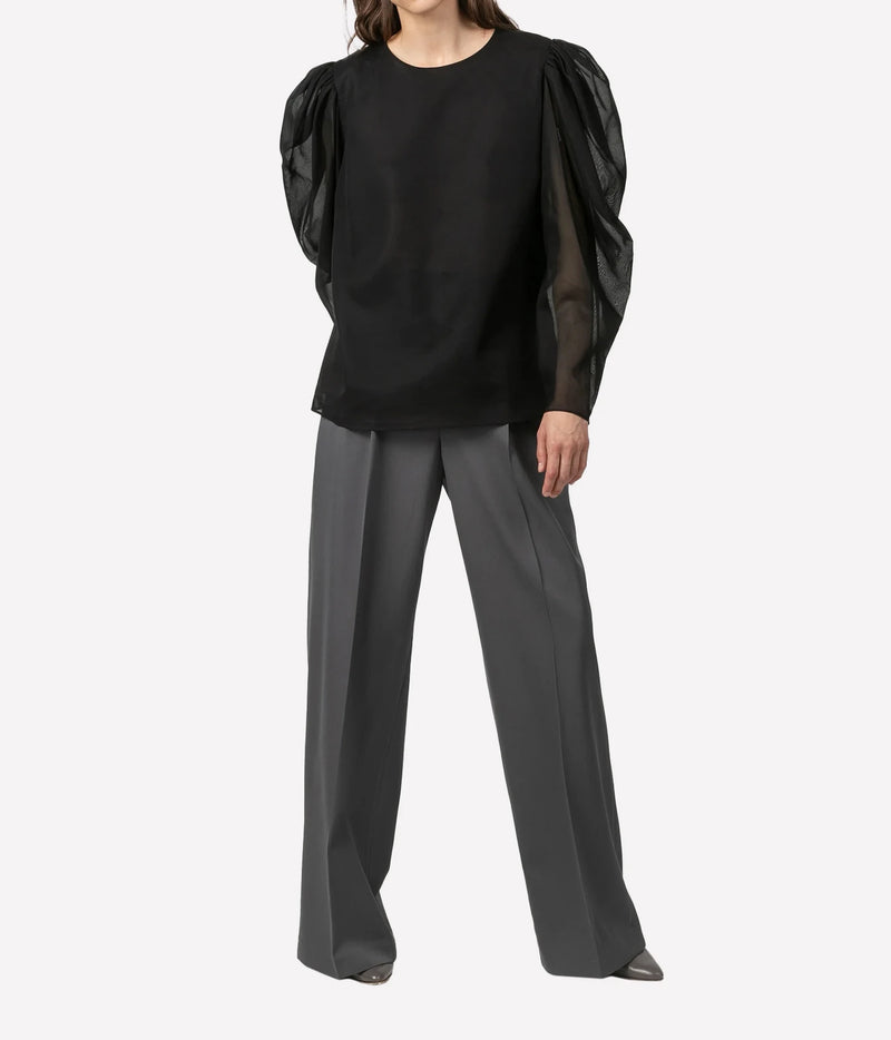 A model wearing a black long-sleeve blouse with dramatic sheer puff sleeves and dark gray wide-leg trousers, photographed against a clean light gray studio background.