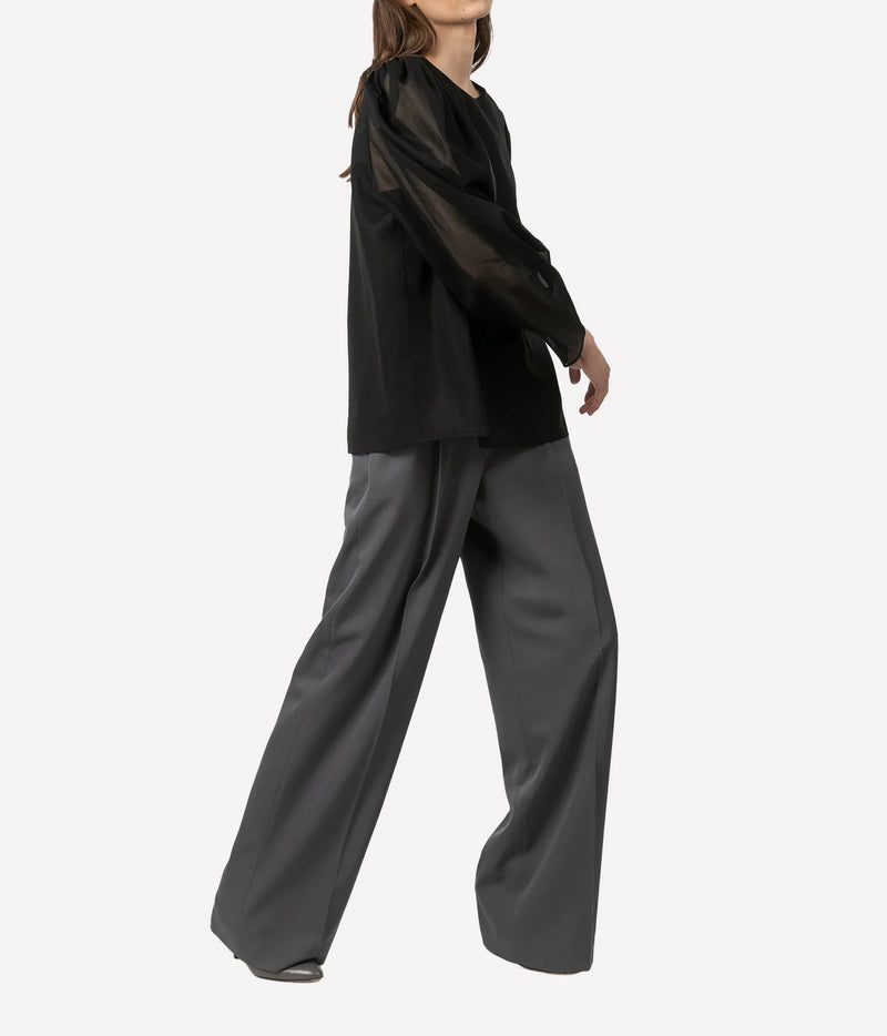 A model wearing a black sheer long-sleeve top and dark gray wide-leg trousers, shown from a side profile against a plain white background.