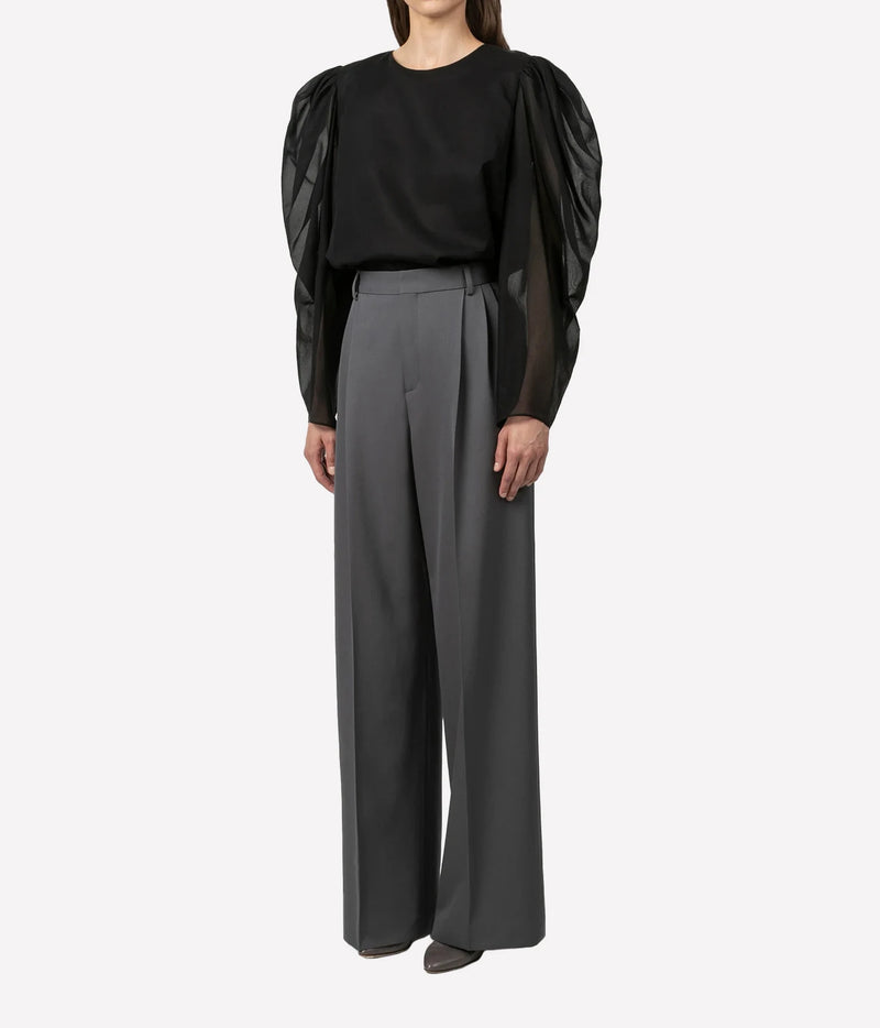 A model wearing a black long-sleeve top with dramatic sheer puff sleeves and high-waisted, wide-leg gray trousers, photographed against a plain light gray studio background.