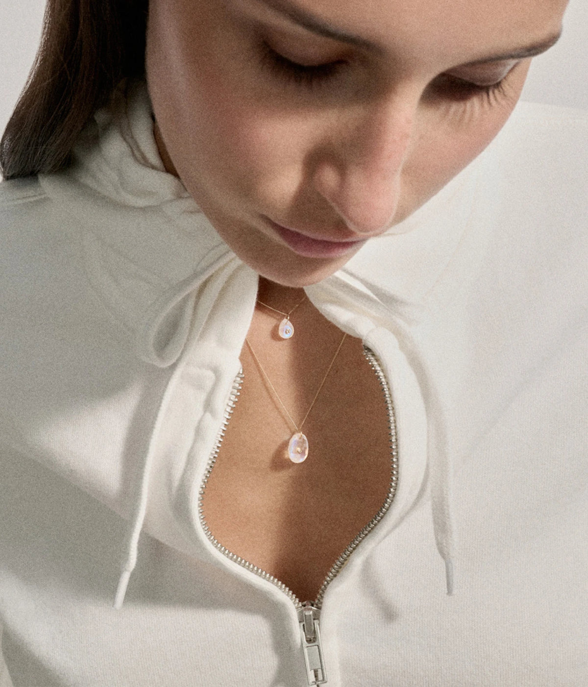 Model looking down wearing the Pascale Monvoisin Orso Moonstone Necklace with a white zip-front top open at the neckline, the moonstone pendant sitting at the centre of the chest