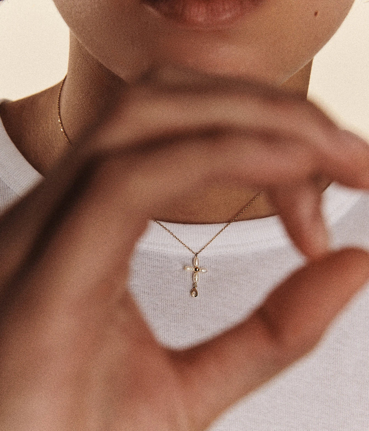 Model wearing the Pascale Monvoisin Palerme White Pearl Necklace with a white top, hands raised to the chin and the pearl cross resting below the collarbone