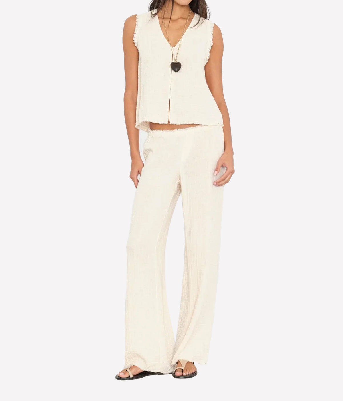 Model wearing the Xirena Joseph Pant in Oysterette with a matching sleeveless button-front vest, a black heart pendant necklace and dark thong sandals, showing the relaxed full-length leg