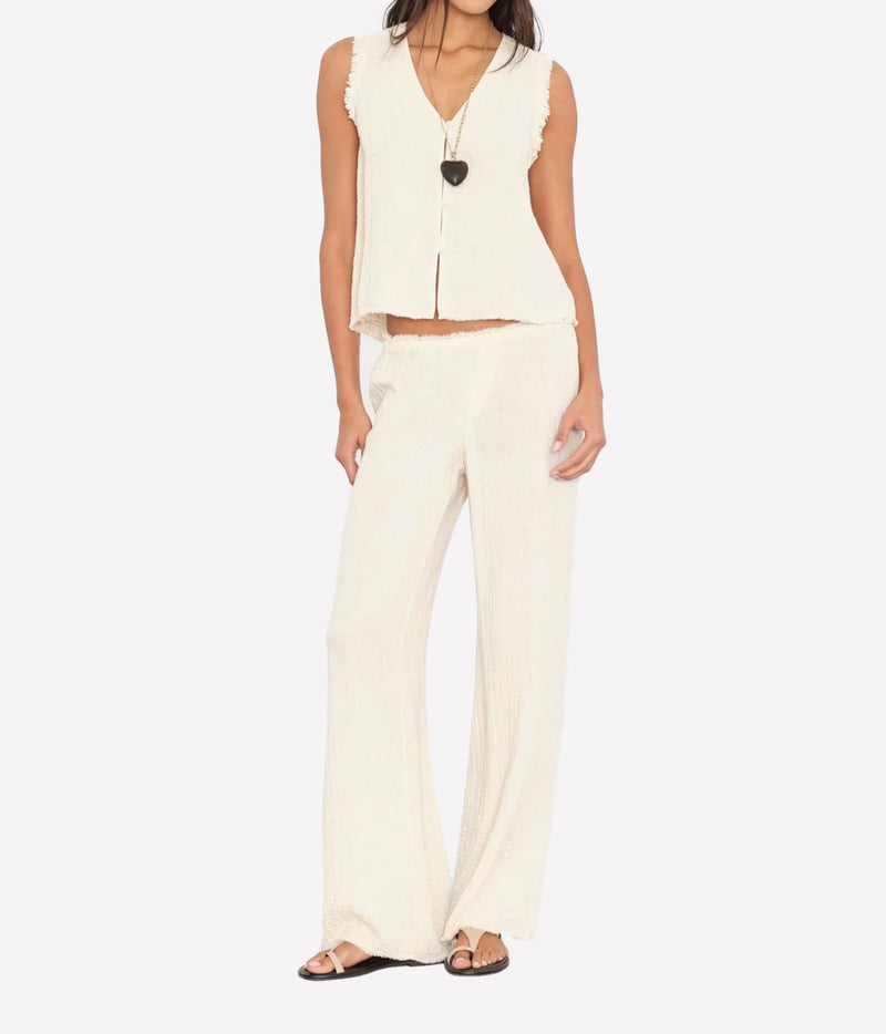Model wearing the Xirena Joseph Pant in Oysterette with a matching sleeveless button-front vest, a black heart pendant necklace and dark thong sandals, showing the relaxed full-length leg