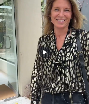 Woman wearing the Kempton & Co. Mia Suede Stud Shoulder Bag in Black on the shoulder, styled with a black and gold print blouse and dark jeans, walking outside a boutique