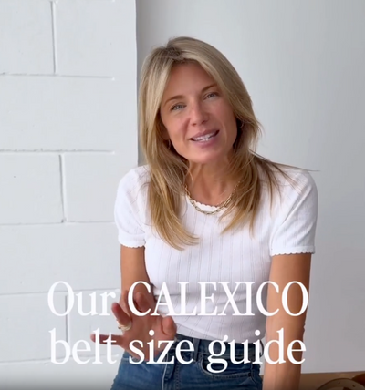 Calexico founder Nicky in a white pointelle tee and blue jeans, hands on her hips, explaining how Calexico belts are sized, with brown, tan and studded leather belts draped beside her