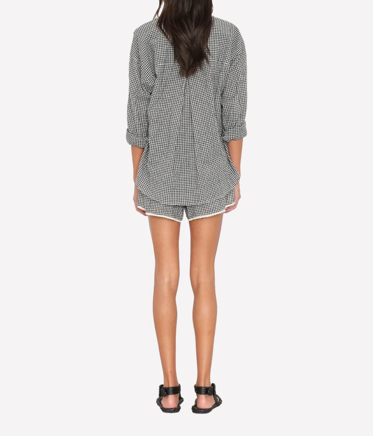 Back view of a model wearing the Xirena Starla Short in Black Sand with an oversized gingham shirt and black strappy sandals, showing the trimmed hem