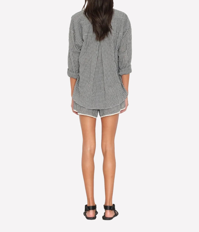Back view of a model wearing the Xirena Starla Short in Black Sand with an oversized gingham shirt and black strappy sandals, showing the trimmed hem