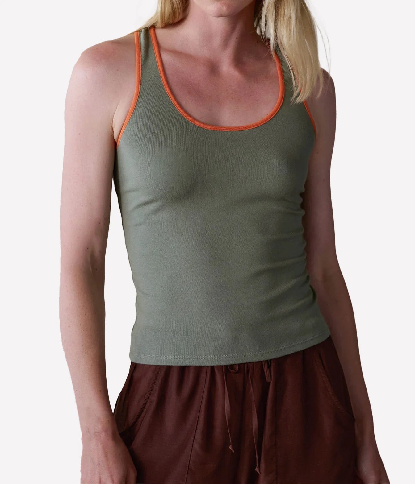 A woman wearing an olive green ribbed tank top with bright orange trim along the scoop neckline, paired with brown drawstring pants, against a plain light gray background.