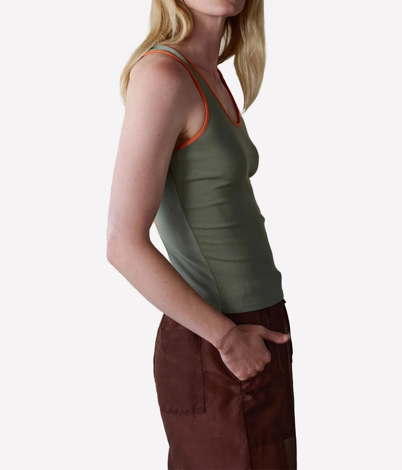 A side-profile view of a woman wearing an olive green ribbed tank top with orange trim and brown pants, set against a plain light gray background.
