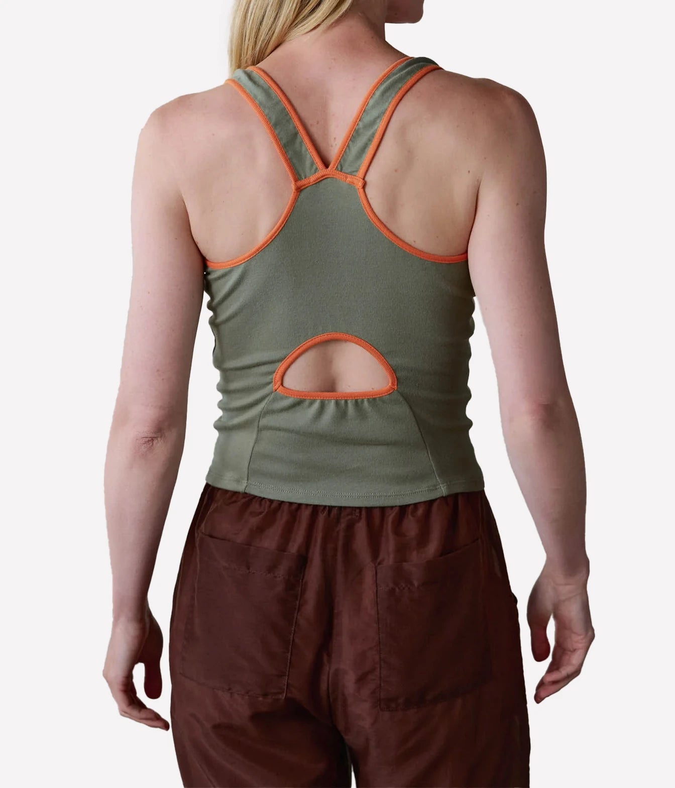A woman shown from the back wearing an olive green racerback tank top with orange trim and a cutout at the lower back, paired with brown pants, against a light gray studio background.