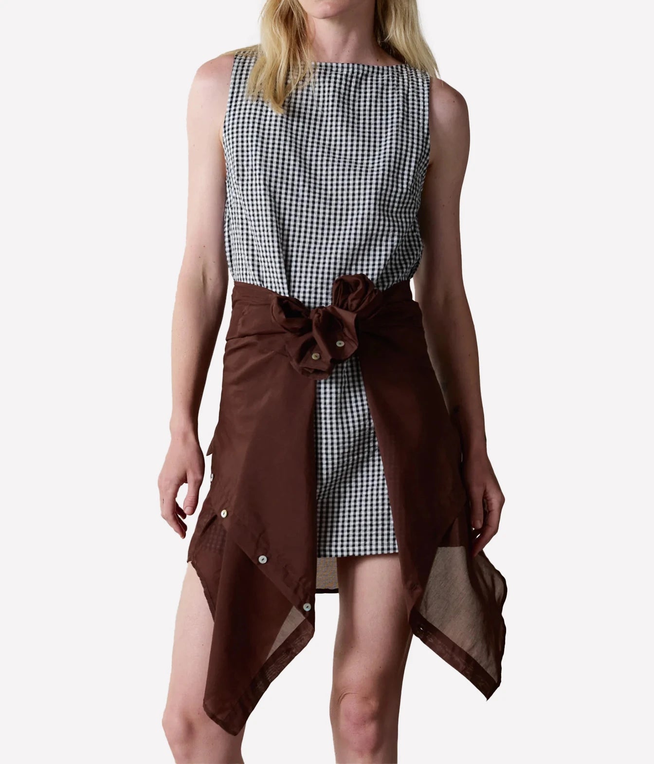 A model wearing a sleeveless black and white gingham dress with a dark brown asymmetrical wrap skirt overlay featuring snap details and a bow at the waist.