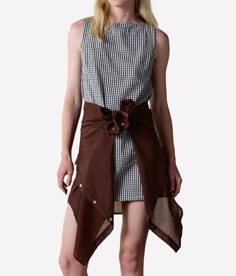 A model wearing a sleeveless black and white gingham dress with a dark brown asymmetrical wrap skirt overlay featuring snap details and a bow at the waist.