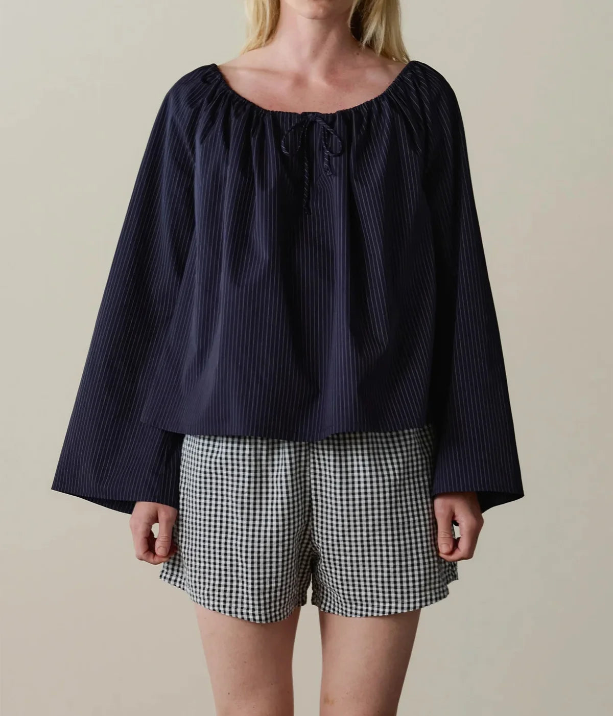 A woman wearing a navy blue off-the-shoulder top with vertical pinstripes and the DONNI. The Cotton Gingham Tap Short in JET in black-and-white gingham