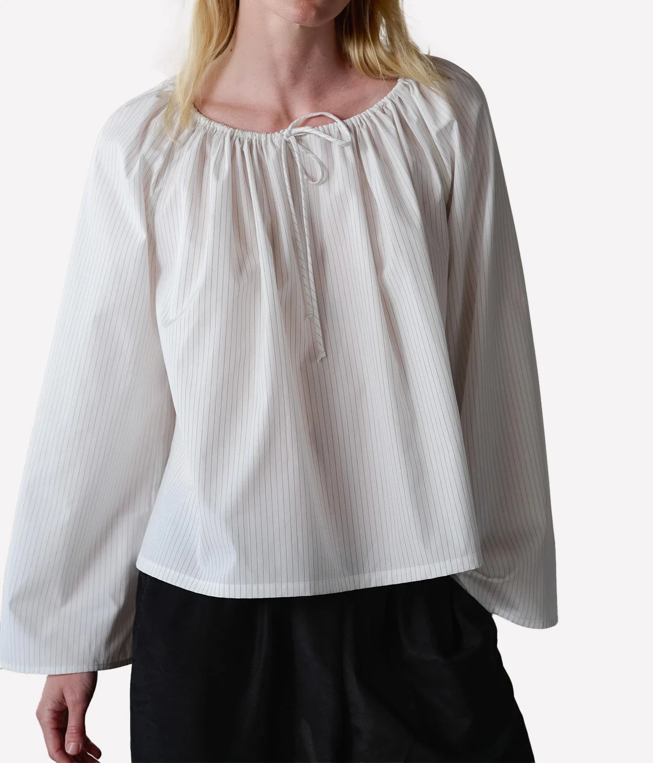 A woman wearing a white long-sleeve blouse with subtle vertical pinstripes, a gathered round neckline, and a front tie detail, paired with black pants against a light gray studio background.