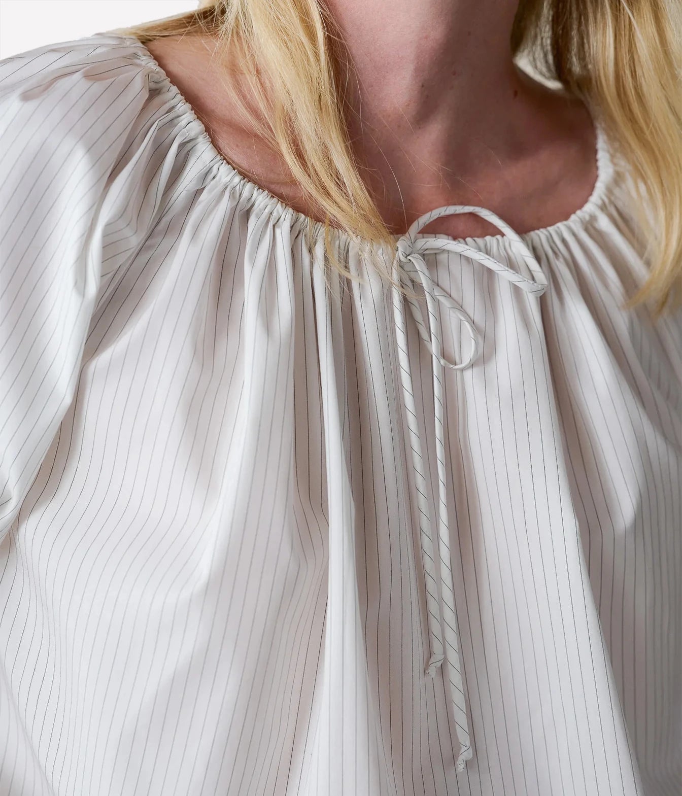 Close-up of a woman wearing a white blouse with thin vertical pinstripes, a gathered round neckline, and a front tie detail.