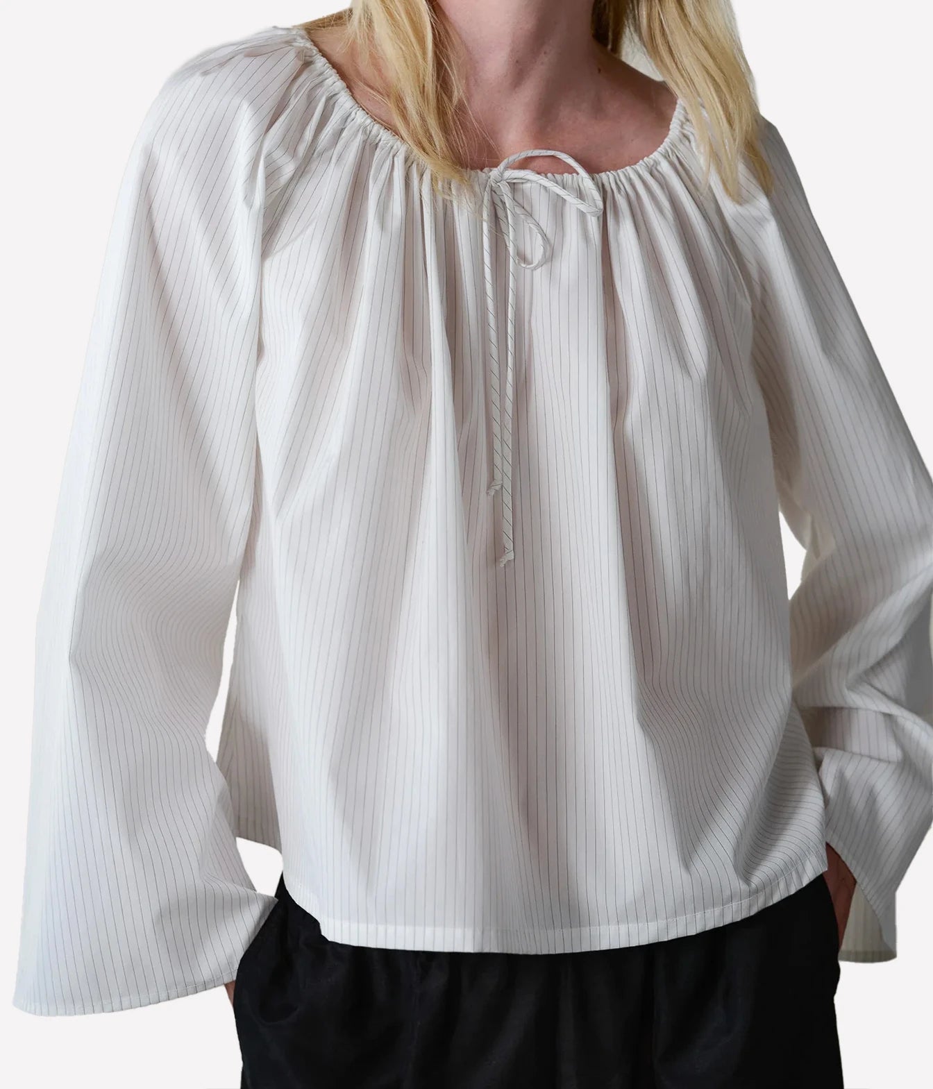 A woman wearing a white long-sleeve blouse with a gathered round neckline and a front tie detail, paired with black bottoms, against a plain light background.