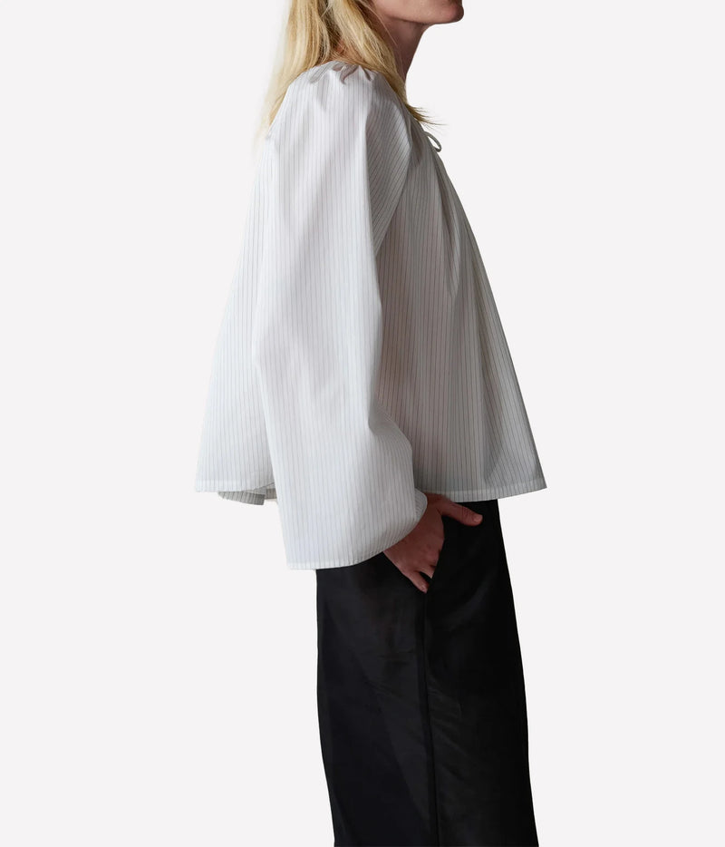 A side-profile view of a model wearing a white long-sleeve blouse with subtle vertical pinstripes and black wide-leg trousers, set against a plain white studio background.