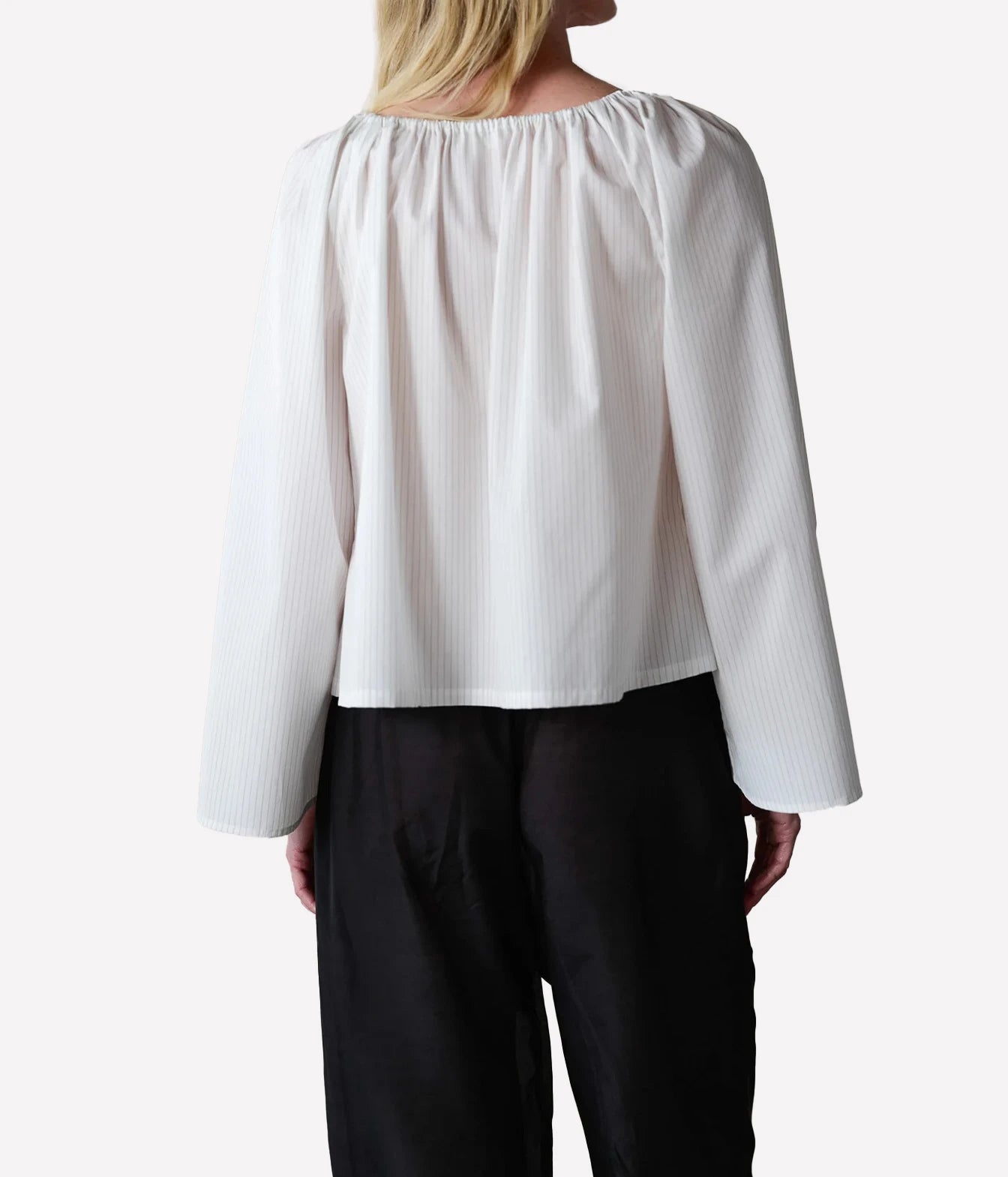 A woman shown from the back wearing a white long-sleeve blouse with a gathered, pleated back and black trousers, against a plain light gray studio background.