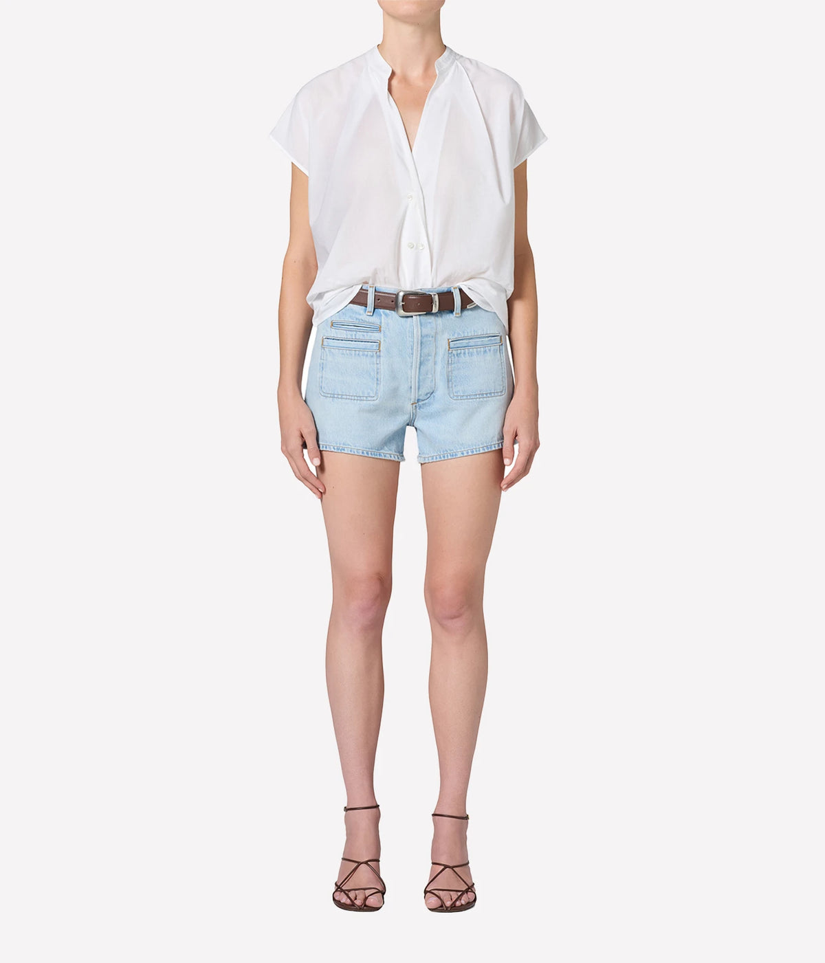 Full-length front view of a model wearing the Citizens Of Humanity Marlow Welt Pocket Short in Lasso with a white short-sleeve blouse tucked in, a brown leather belt and dark strappy heeled sandals