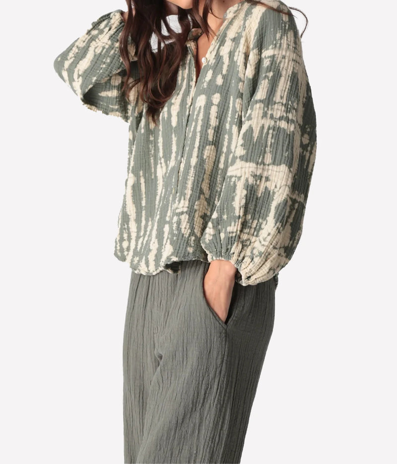 Side angle of a model with one hand raised and the other in her pocket in the Electric Rose Winnie Horizon Shirt in Moss Ivory, showing the blouson fit and elasticated cuffs, with moss green crinkle-textured pants