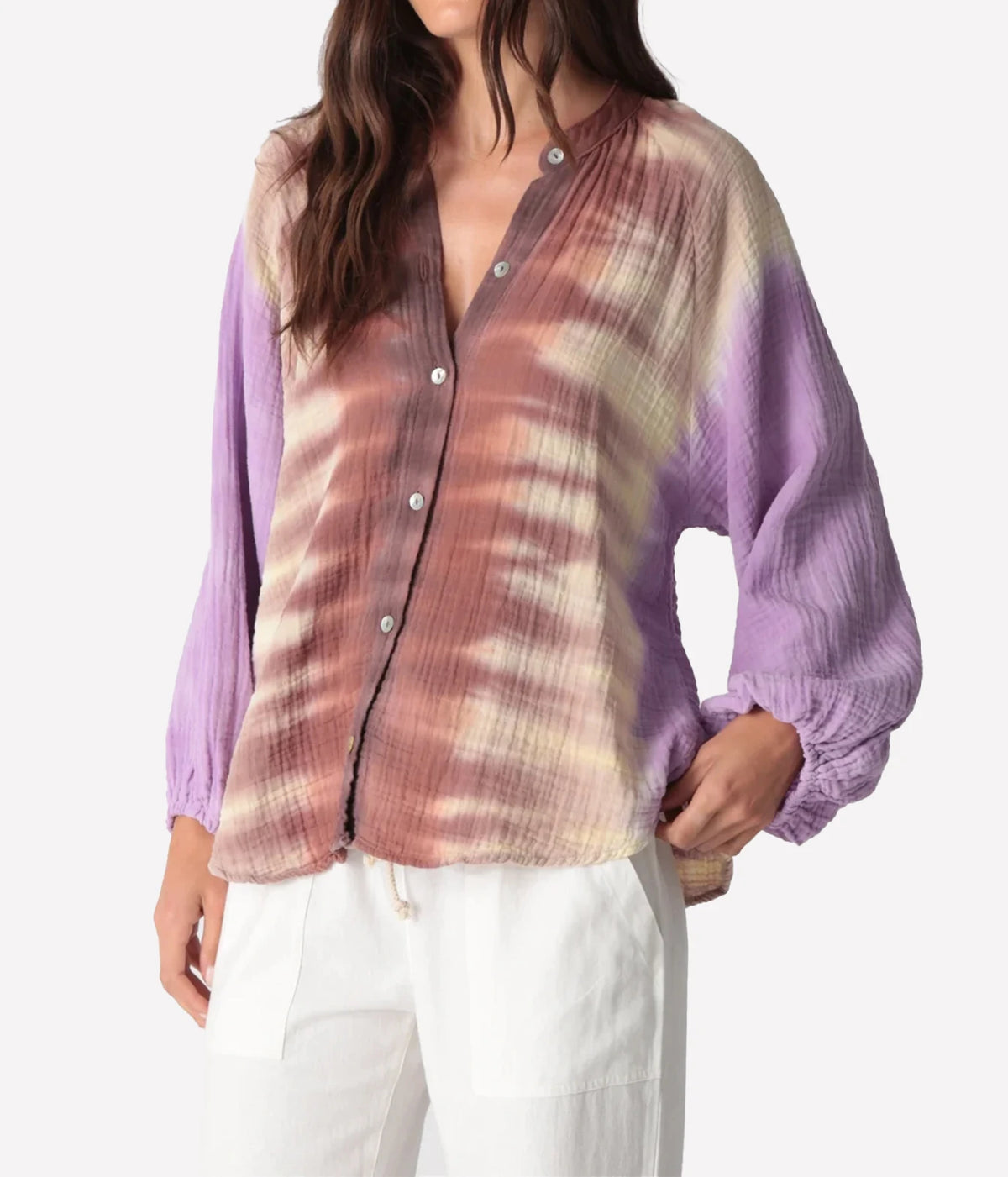 Close front view of a model with a hand on her hip in the Electric Rose Winnie Fade Shirt in Lavender Sepia, showing the button front, crinkled cotton gauze and elasticated cuffs, with white drawstring trousers