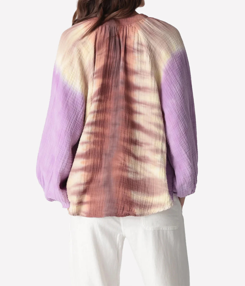 Back view of a model wearing the Electric Rose Winnie Fade Shirt in Lavender Sepia, showing the gathered yoke, sepia tie-dye down the back and lavender sleeves, with white trousers