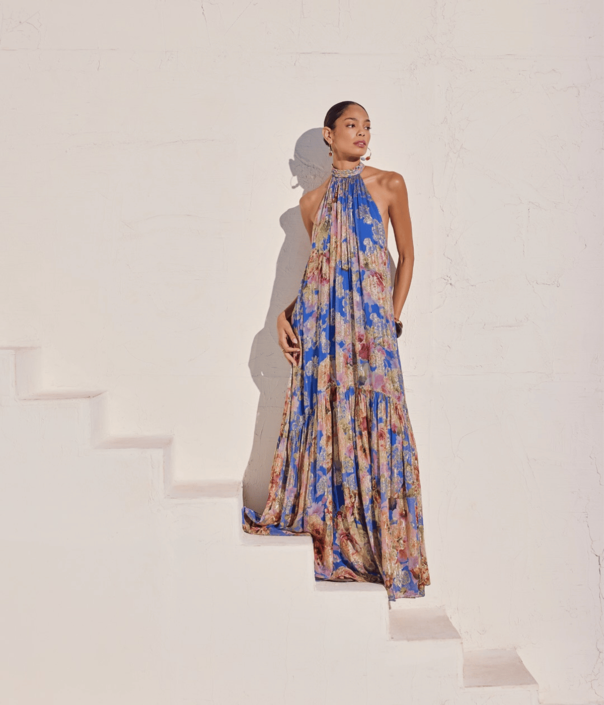 A model wearing a halter-neck, tiered maxi dress with a vibrant blue, pink, and beige floral pattern, posing on white stairs against a textured white wall.