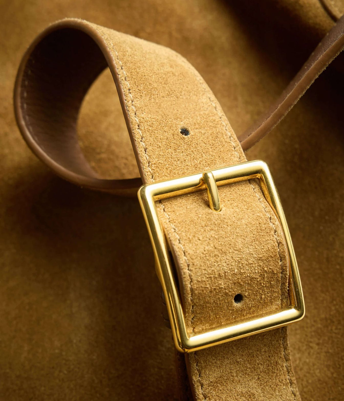 A close-up of a tan suede belt with a polished gold-tone metal buckle, resting on a matching brown suede background.