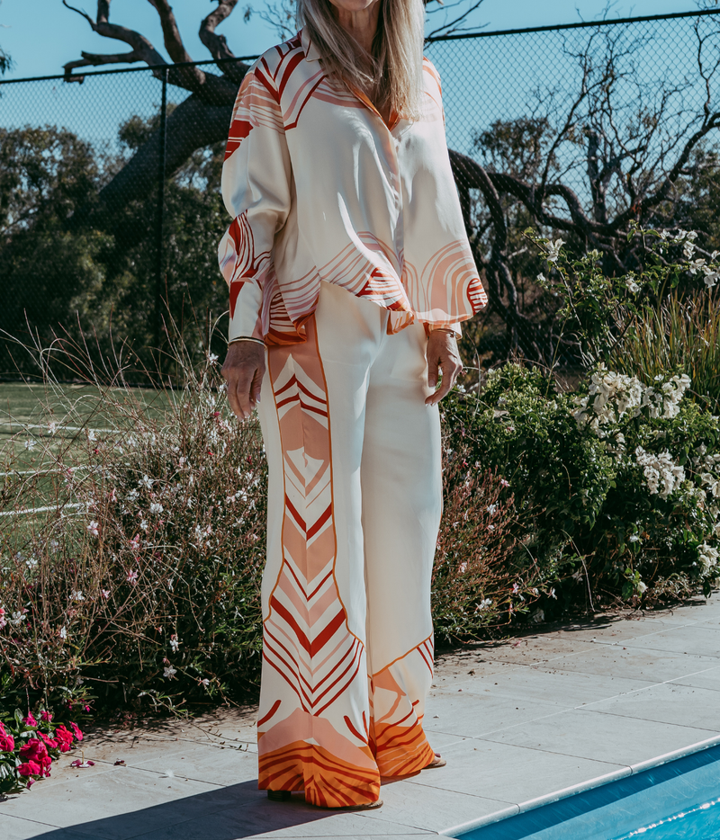 A woman wearing a matching two-piece lounge set with a cream base and orange geometric patterns, standing outdoors next to a swimming pool.