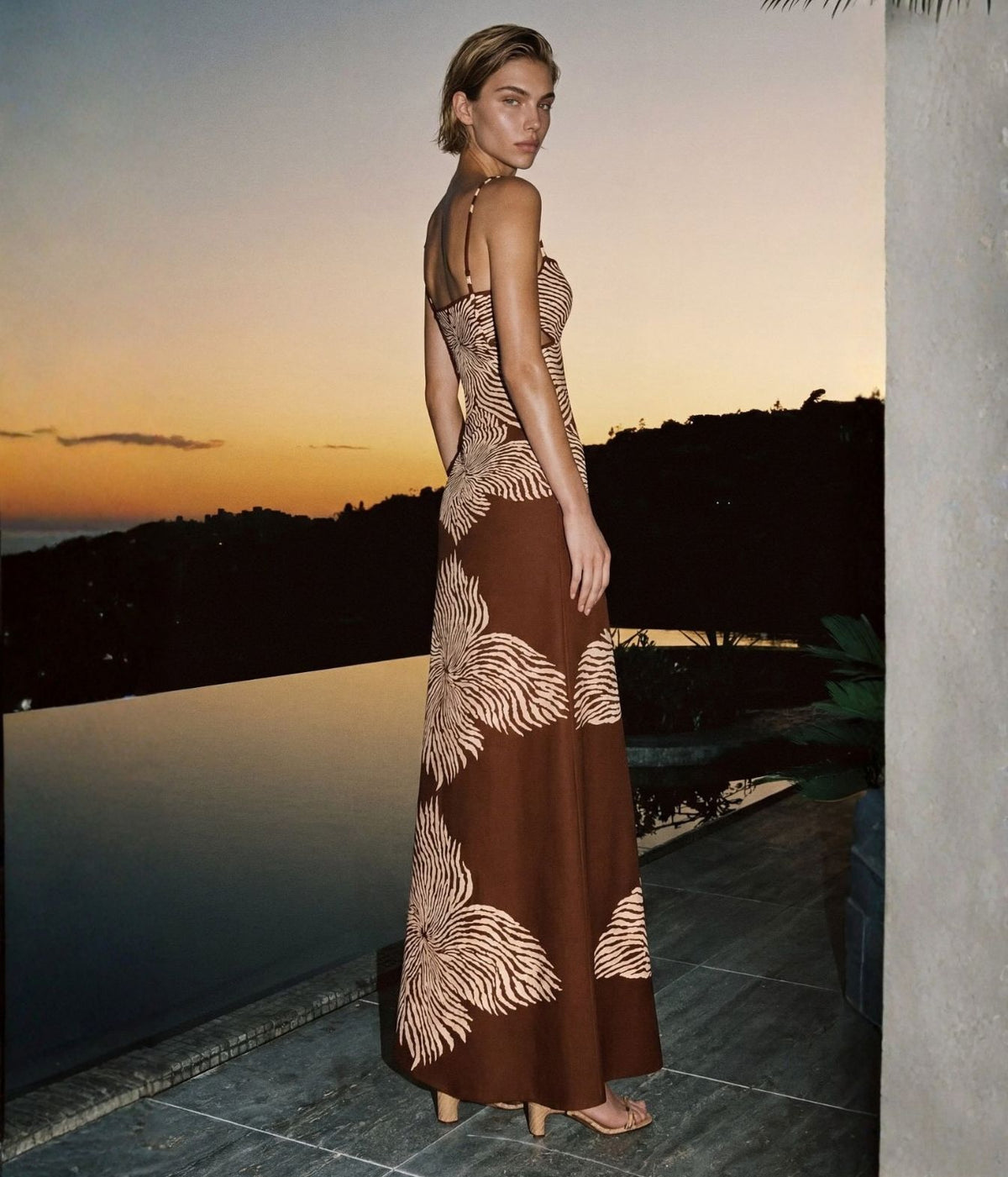 Campaign image of woman wearing Alexis Rayana Dress In Brown Blossom, standing by a pool at sunset with raffia look heels