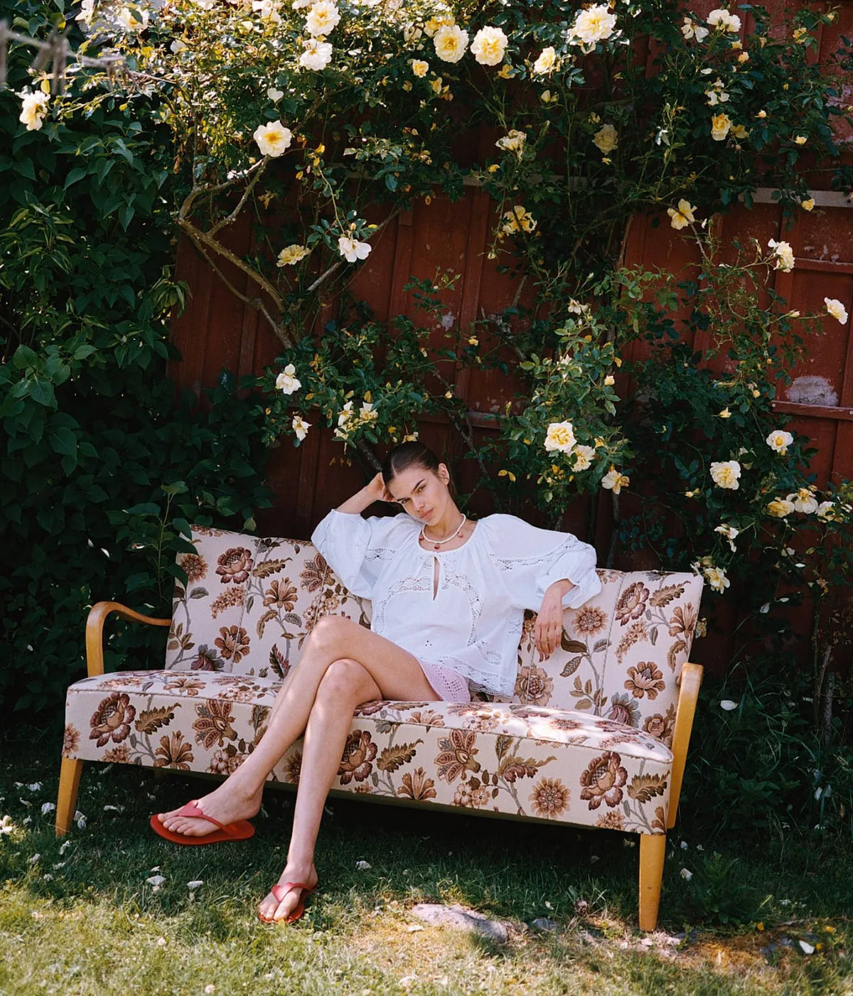 Model reclining in the Sissel Edelbo Agathe Top In Ecru with pale pink skirt, showing the crochet lace insets and embroidery across the front