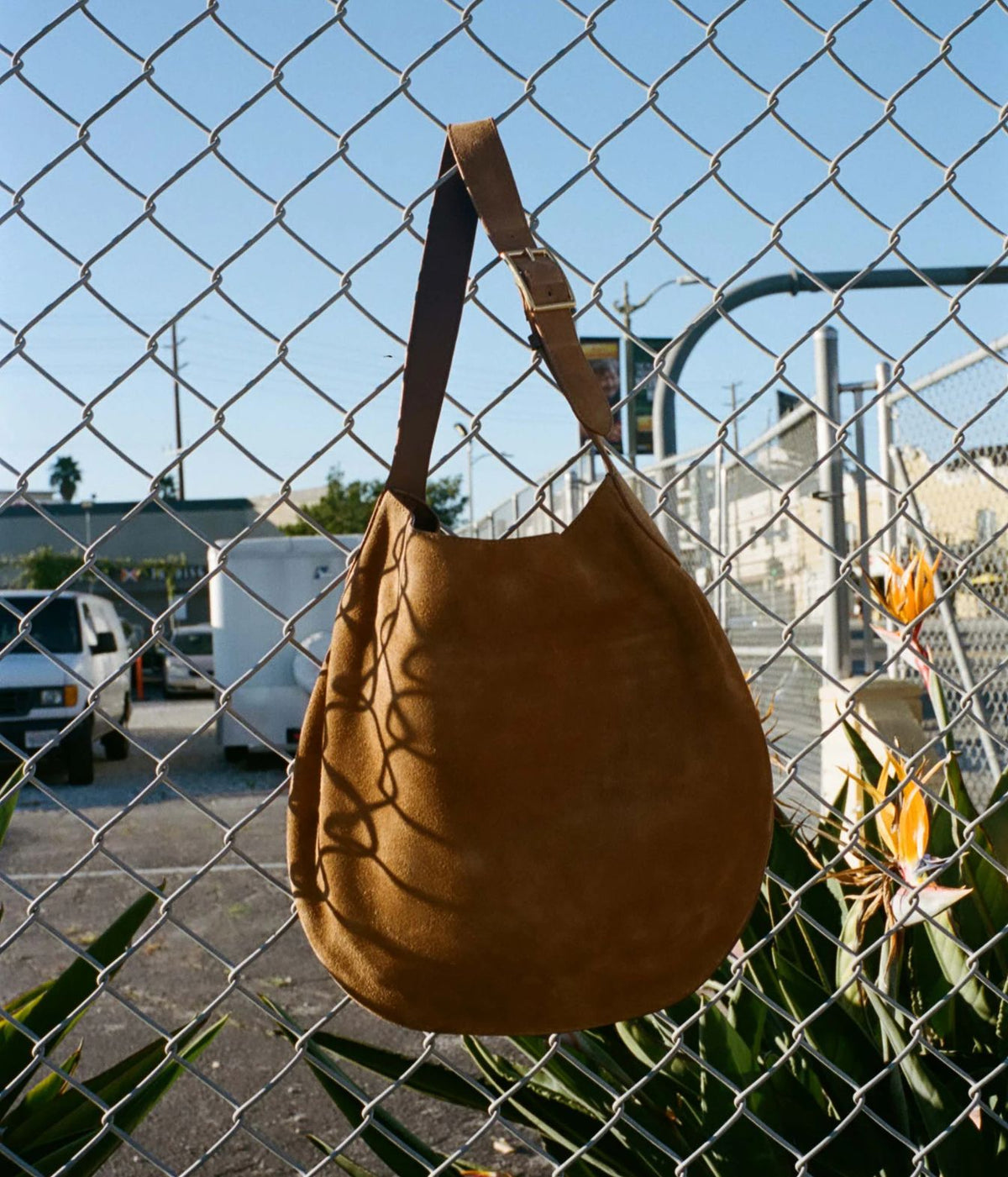 A brown suede shoulder bag hanging in front of a chain-link fence outdoors, with a blue sky and industrial background visible behind it.
