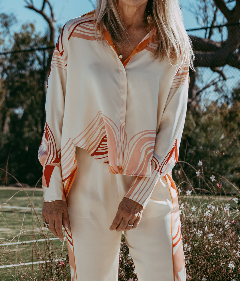 A woman wearing a cream-coloured long-sleeve button-up shirt and matching pants with orange and peach abstract wavy stripe patterns, standing outdoors in a grassy field.